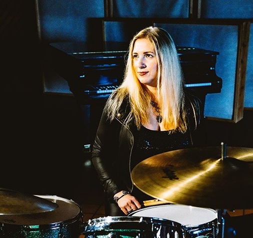 Karen Biller looks off and smiles while sitting at a drum set.