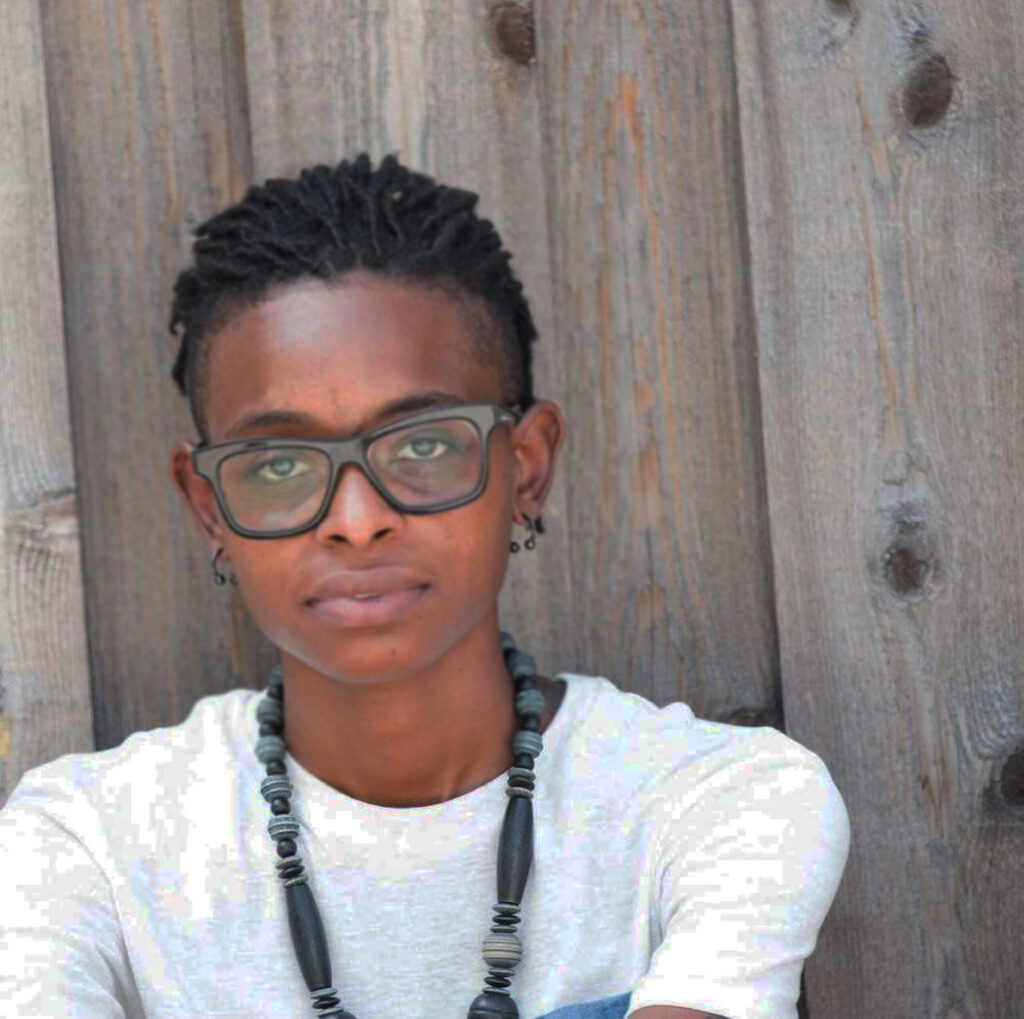 Brianne Sargent, wearing glasses, a white t-shirt, and a necklace, sits against a wooden fence.